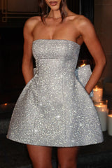 Close-up of a woman in a shimmering silver strapless dress with a flared skirt and a prominent bow at the back, styled for an evening event.