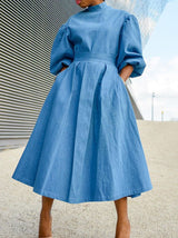 Woman wearing a blue denim midi dress with puffed sleeves and a belted waist, standing outdoors.