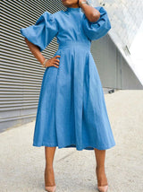 Woman in a blue denim midi dress with puffed sleeves, hands on hips, outdoors.