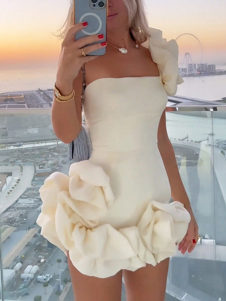 Woman in a white one-shoulder mini dress with ruffled details, standing on a balcony with a city view at sunset.