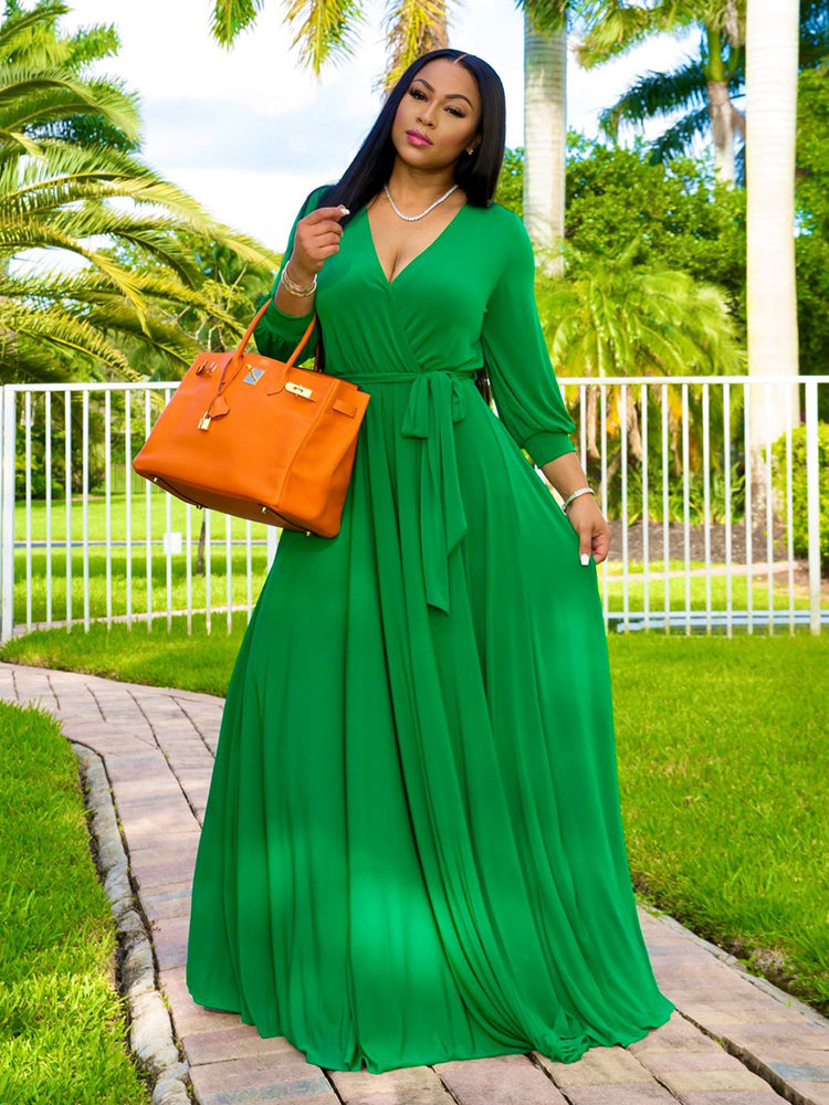 Woman wearing a long green wrap dress with a V-neck and long sleeves, holding an orange handbag, standing outdoors with palm trees in the background.