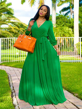 Woman wearing a long green wrap dress with a V-neck and long sleeves, holding an orange handbag, standing outdoors with palm trees in the background.