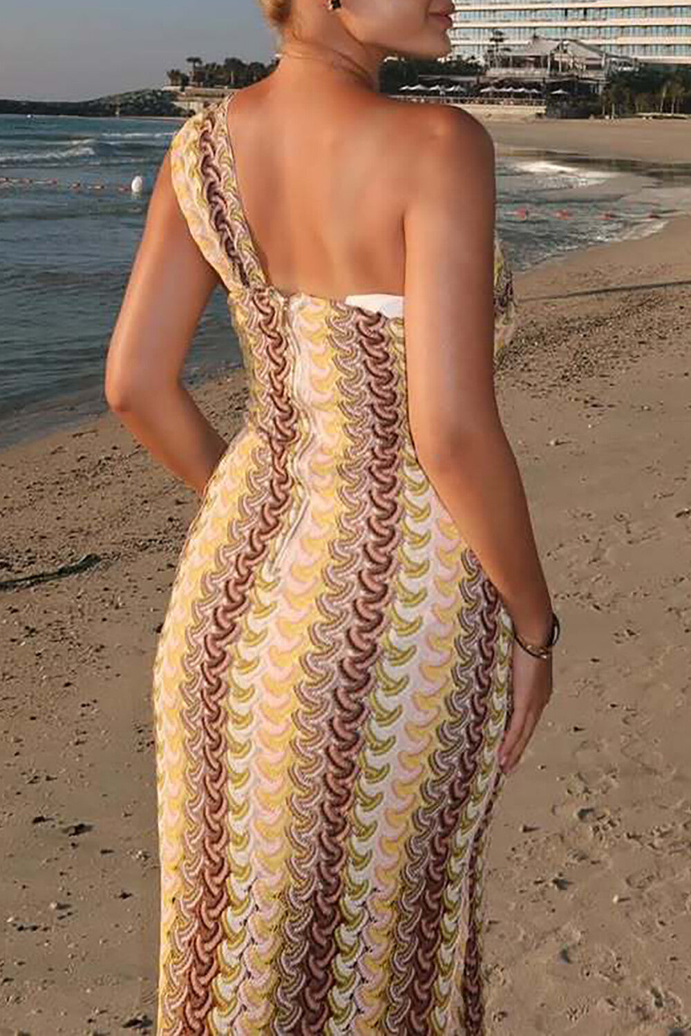 Back view of a woman wearing a yellow and brown patterned one-shoulder knit dress on a beach.