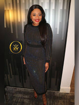 Woman in a black long-sleeved midi dress with vertical rhinestone embellishments, standing in front of a wall with vertical gold and black stripes. She is wearing red lipstick and gold earrings.
