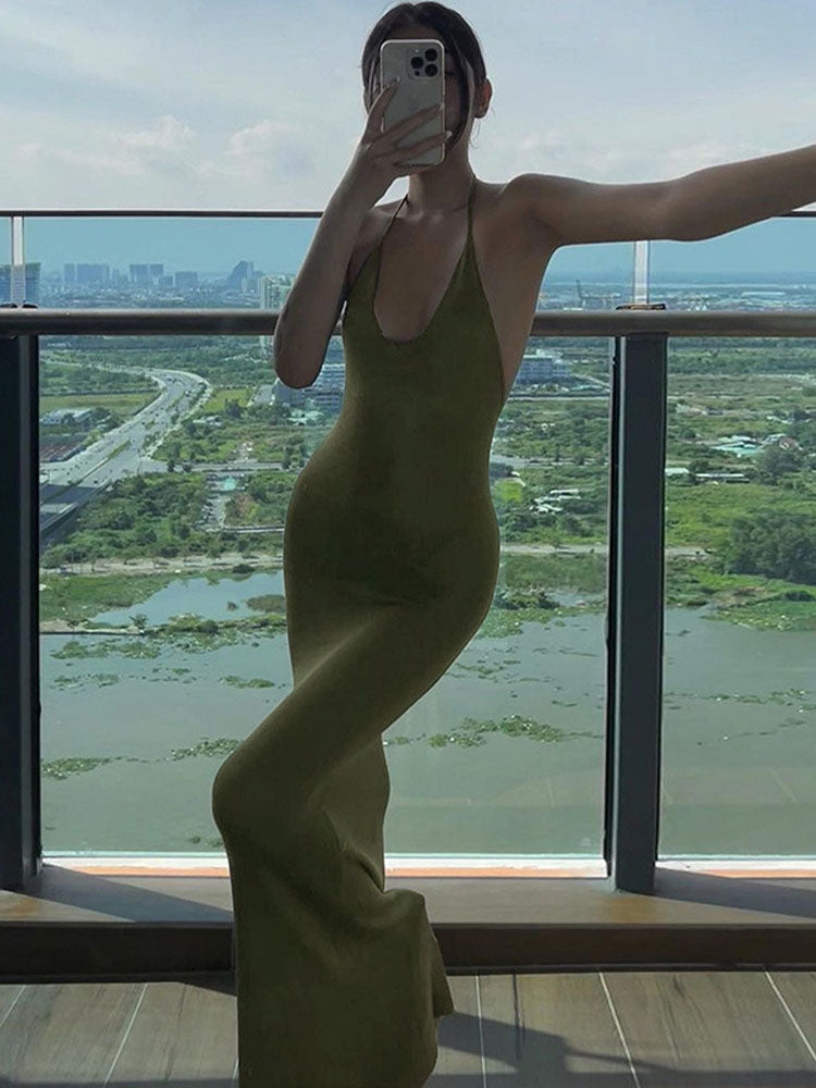 woman wearing an olive green fitted maxi dress with a halter neckline, posing on a balcony with a city view