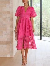 Full body shot of a woman in a hot pink midi dress with a sheer grid overlay, puffed short sleeves, and a button-down front, paired with nude heels and a light pink handbag.