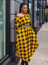 Close-up of a woman in a yellow and black plaid off-the-shoulder midi dress with a gold belt, leaning against a building.