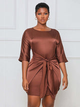 Close-up of a woman wearing a brown satin mini dress with a tie waist and round neckline from the front.
