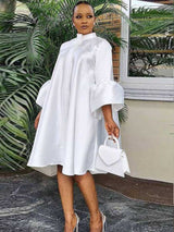 Woman wearing a white A-line dress with bell sleeves and a high neckline, holding a white handbag.