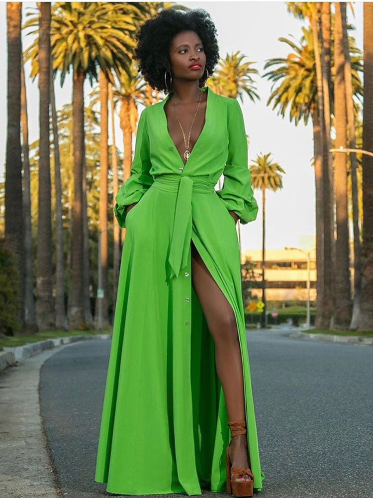 Woman in a bright green maxi dress with a plunging neckline, belted waist, and a thigh-high slit, posing on a street lined with palm trees.