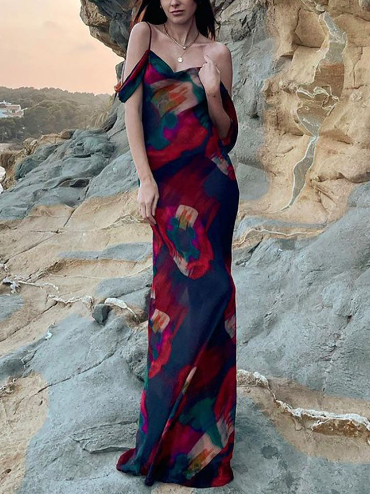 Woman wearing a flowing abstract floral print maxi dress with cold shoulder sleeves standing on rocky terrain near the ocean.