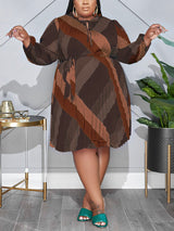 Plus-size woman wearing a brown, long-sleeved, pleated A-line dress with a geometric print and tie-waist.