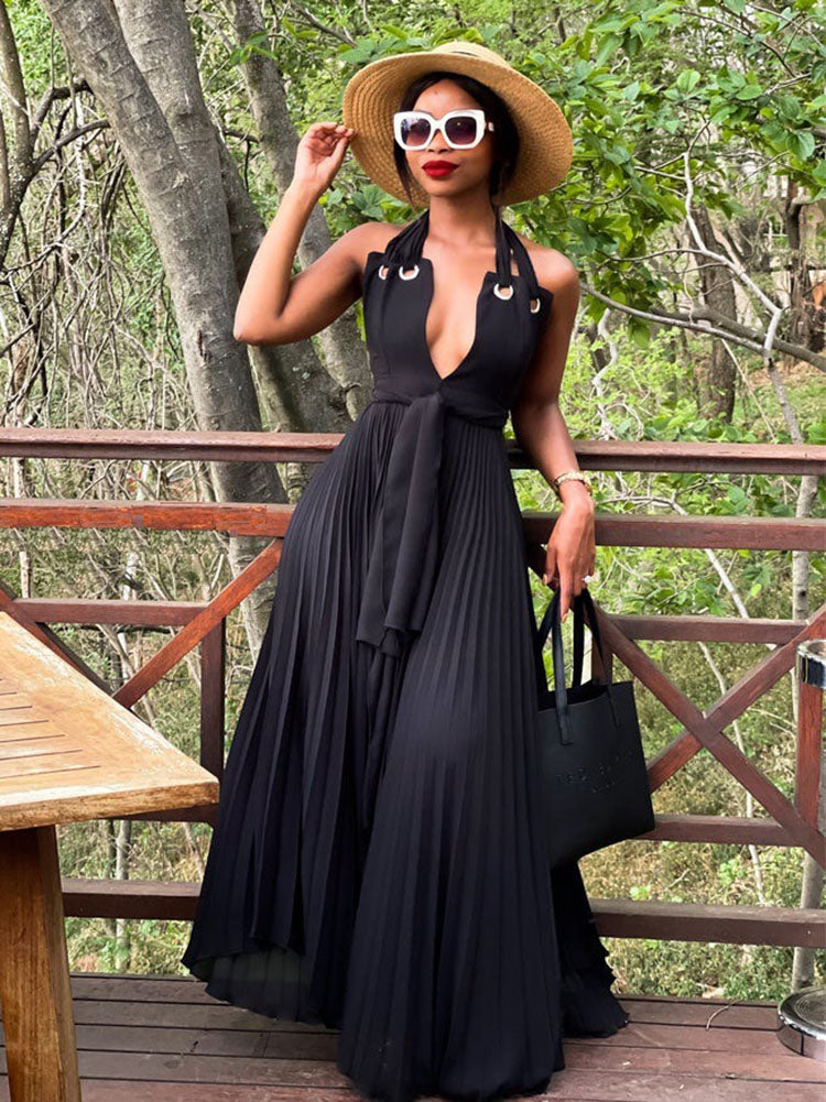 Woman wearing a black halter maxi dress with a deep V-neck and pleated skirt, accessorized with a straw hat and sunglasses, standing on a wooden deck outdoors.