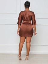 Woman wearing a brown satin mini dress with a tie waist and round neckline from the back.