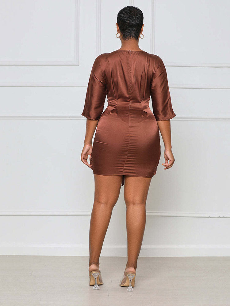 Woman wearing a brown satin mini dress with a tie waist and round neckline from the back.