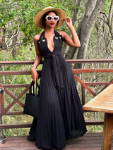 Full body shot of a woman in a black halter maxi dress with a pleated skirt, holding a black tote bag.
