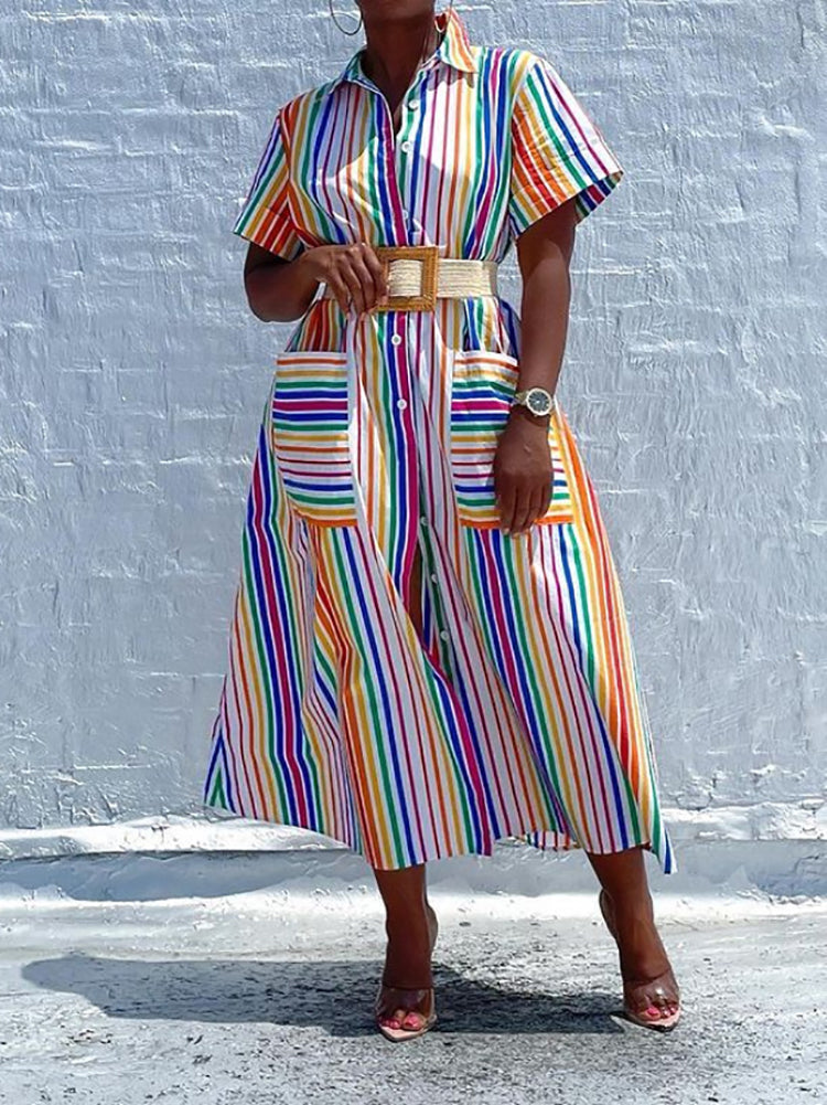 Woman wearing a belted rainbow striped shirt dress with a high slit and pockets, standing against a white brick wall.