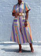 Woman wearing a belted rainbow striped shirt dress with a high slit and pockets, standing against a white brick wall.