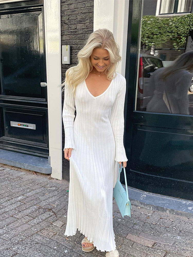 Woman with blonde hair wearing a white ribbed knit maxi dress with flared cuffs, smiling and carrying a light blue handbag.