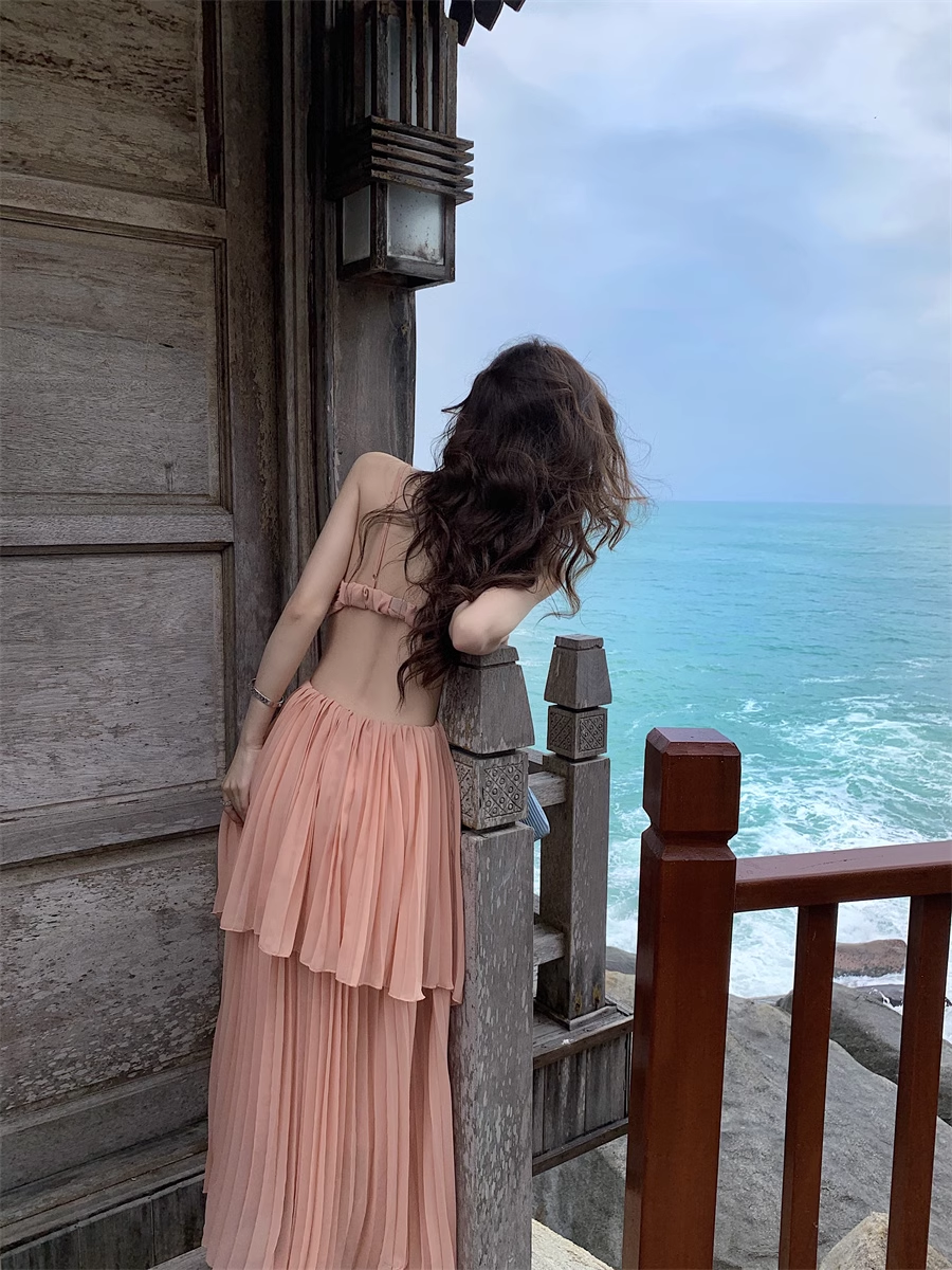 Woman wearing a peach halter neck maxi dress with a cut-out and pleated tiered skirt, leaning against a railing with her back to the camera, overlooking the ocean.
