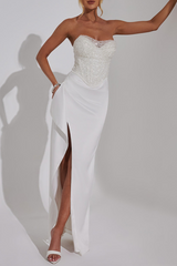 Woman wearing a white strapless evening gown with a high slit and embellished bodice against a white background