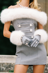 Woman wearing a grey off-the-shoulder mini dress with white faux fur trim and cuffs, grey gloves, and a diamond necklace, hands clasped.