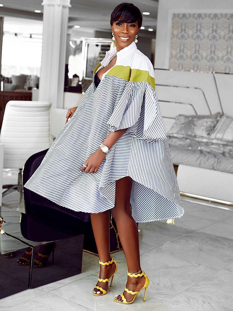 Full-body shot of a woman in a voluminous, striped shirt dress with a green accent, showcasing the asymmetrical hemline.