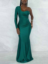 Woman wearing a floor-length, one-shoulder green dress with a long sleeve on the left side and a mermaid hem, standing against a marbled background.