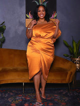 Woman adjusting the fabric of her orange satin one-shoulder dress with a draped bodice and tulip hem skirt, standing in front of a mustard yellow couch.