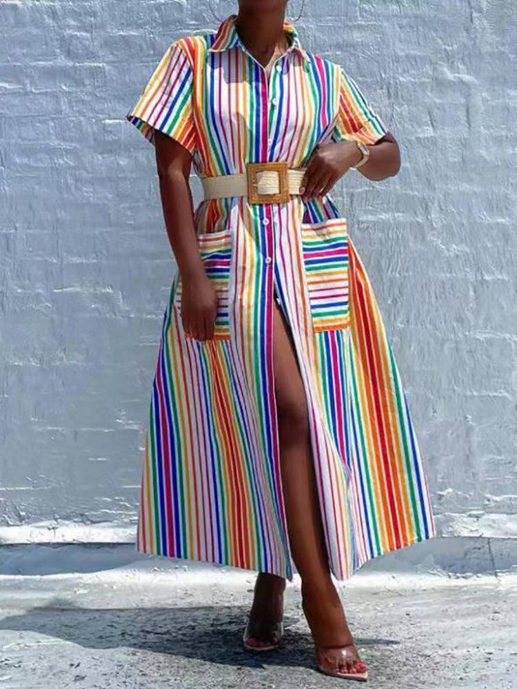 Woman wearing a rainbow striped shirt dress with a high slit and pockets, cinched with a belt, standing against a white brick wall.