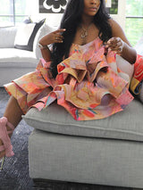 Woman sitting on a sofa, wearing a colorful tiered ruffle dress and pink strappy heels.