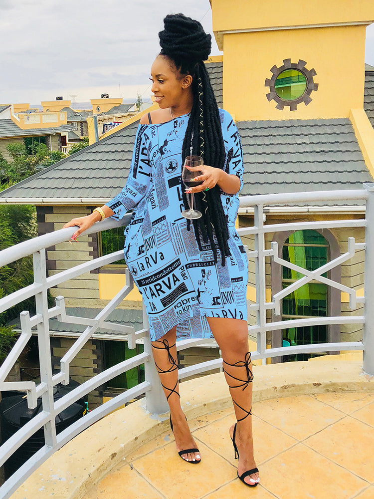 Woman wearing a light blue newspaper print t-shirt dress with black strappy heels, standing on a balcony.