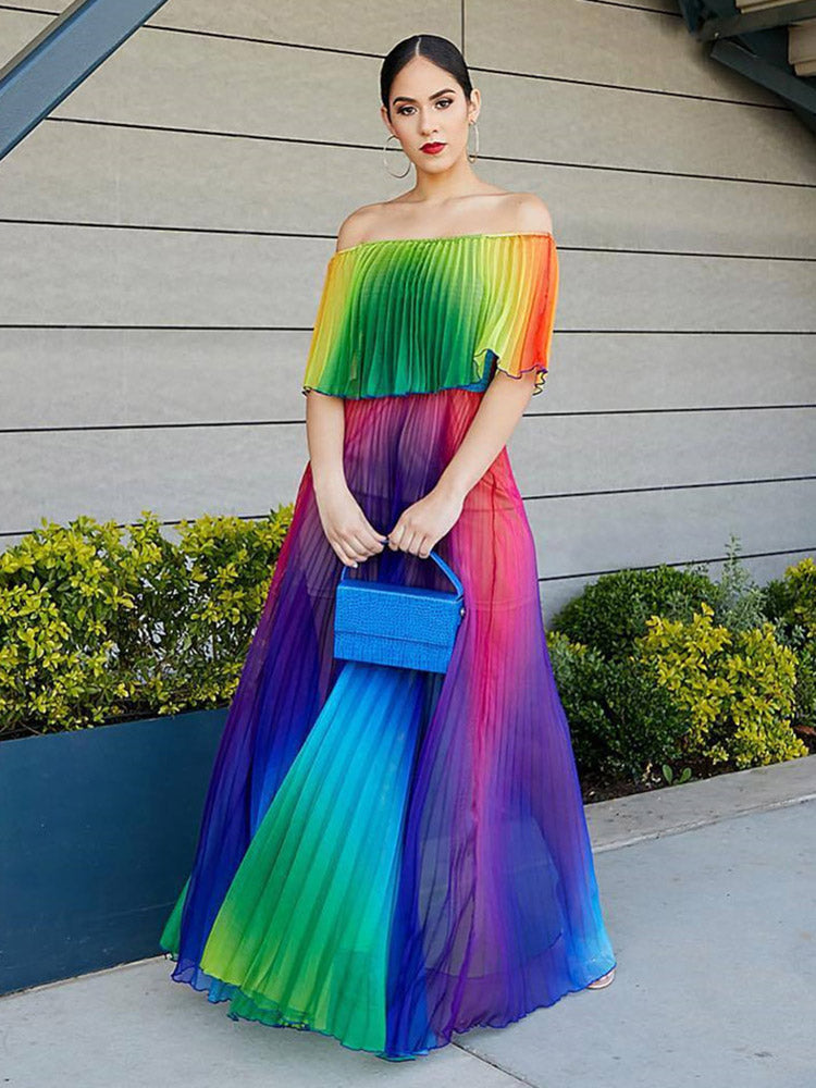 Woman wearing a rainbow gradient off-the-shoulder pleated maxi dress and holding a blue clutch, standing outdoors.
