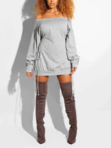 Woman wearing a light grey off-the-shoulder mini dress with drawstring details on the sides and long sleeves, paired with knee-high boots.