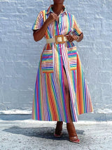 Woman wearing a belted rainbow striped shirt dress with a high slit, standing against a white brick wall.