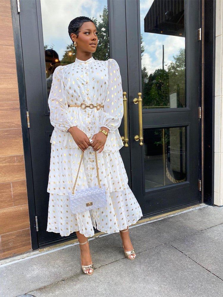 Woman wearing a white tiered midi dress with gold polka dots, a gold belt, and gold heels, standing in front of a black door.