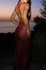 back view of a model wearing a long, sheer, red sequined halter dress at dusk with a body of water in the background