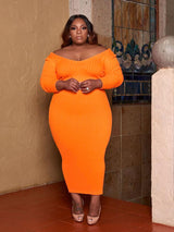 Woman in an orange ribbed knit midi dress with long sleeves and an off-the-shoulder V-neckline, standing with hands clasped.