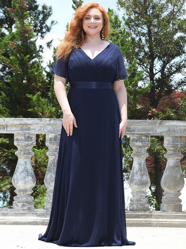 Woman in a navy blue floor-length chiffon dress with flutter sleeves and a V-neckline, standing outdoors.