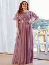 Woman with auburn hair wearing a dusty rose A-line maxi dress with embroidered flutter sleeves and a satin waistband, posing indoors.