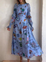 Woman wearing a light blue long-sleeved maxi dress with a graphic print and cut-out waist detail, standing in front of a white wall.