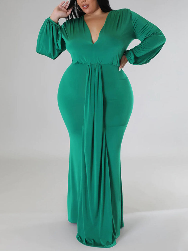 Woman wearing a green, floor-length, long-sleeved gown with a V-neckline and draped skirt, posing with hands on hips
