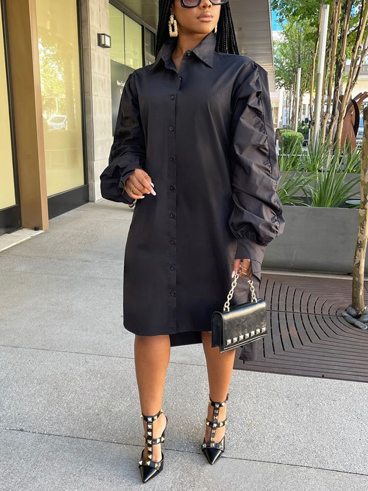 Woman wearing a black collared shirt dress with long ruffled sleeves and an asymmetrical hemline, holding a black clutch and wearing black studded heels.