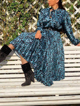 Woman sitting on a white bench wearing a blue leopard print midi dress with a bow neckline and pleated skirt, paired with black embellished boots.
