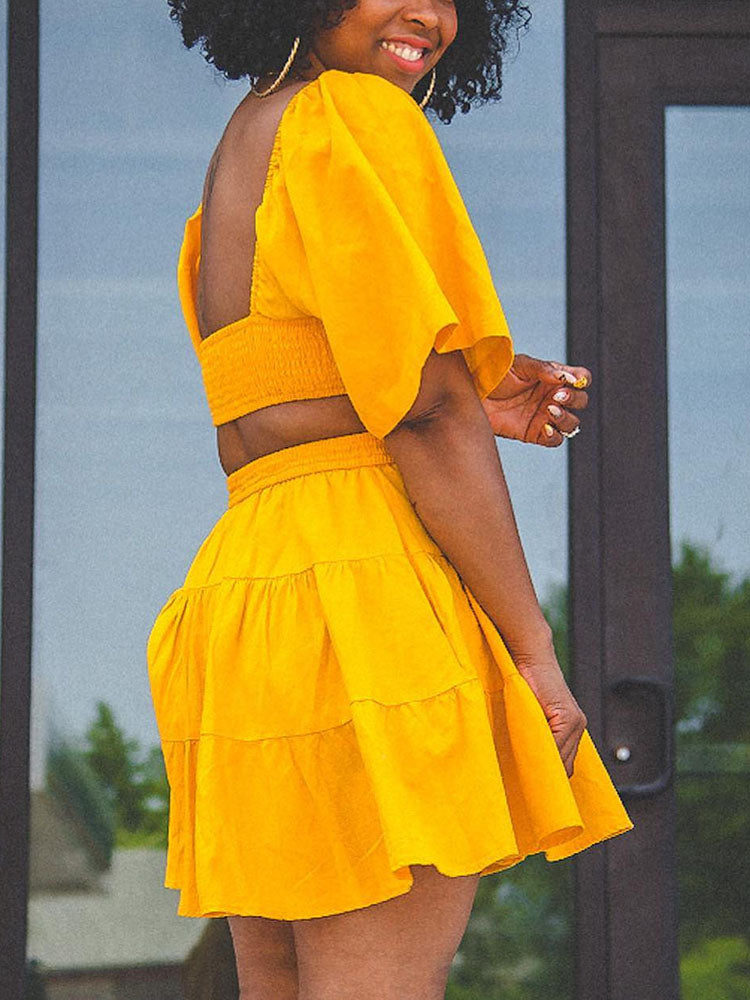 Back view of a woman in a yellow two-piece set with puff sleeves and a tiered skirt.