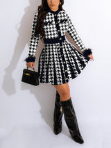 Full-body shot of a woman in a black and white houndstooth knit dress with feather trim cuffs and knee-high boots.