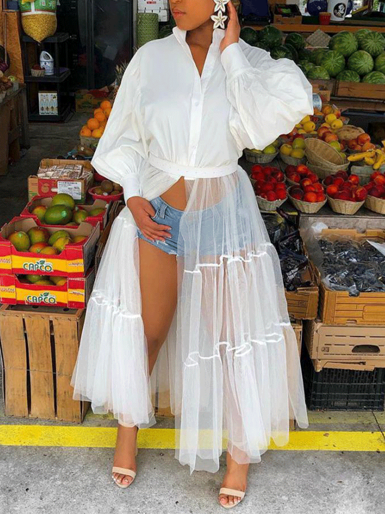 Woman wearing a white button-down shirt dress with a sheer white tiered tulle skirt overlay and denim shorts.