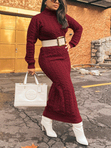 Woman posing in a burgundy cable knit turtleneck maxi dress with a white belt and white boots, holding a white handbag.
