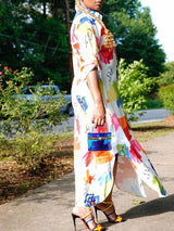 Side view of a woman wearing a long, white, collared shirt dress with a colorful abstract print. The dress has a button-down front, rolled-up sleeves, and a high-low hemline with side slits. She is holding a small blue handbag and wearing colorful high heels.