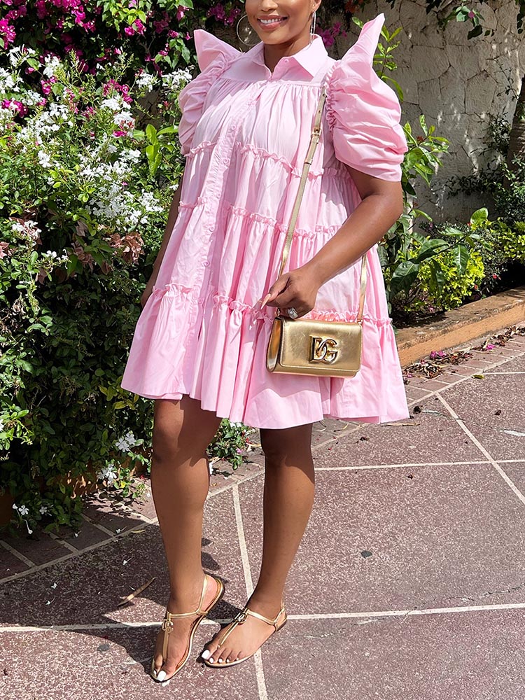 Woman in a pink tiered puff sleeve collared dress, holding a gold handbag and wearing gold flat sandals.